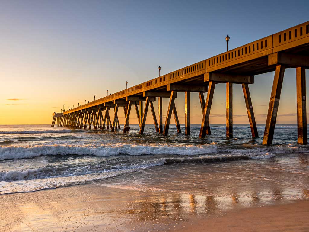 A long pier stretches over the ocean with small waves breaking on the sandy shore at sunset under a clear sky and warm golden light.