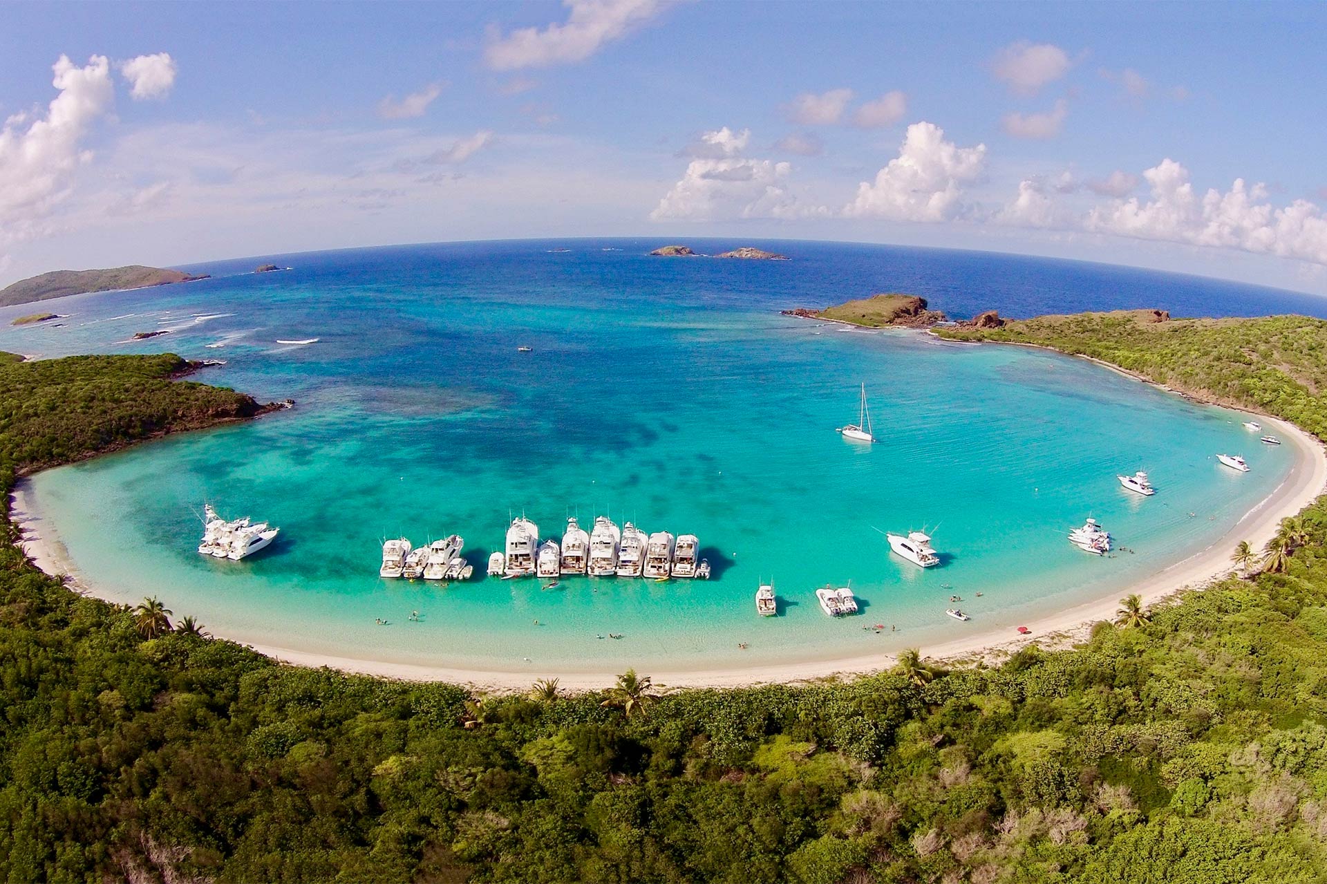 An aerial view of a tropical bay shows white sand, turquoise water, and anchored boats enjoying Memorial Day boating, with lush green vegetation surrounding the shoreline.