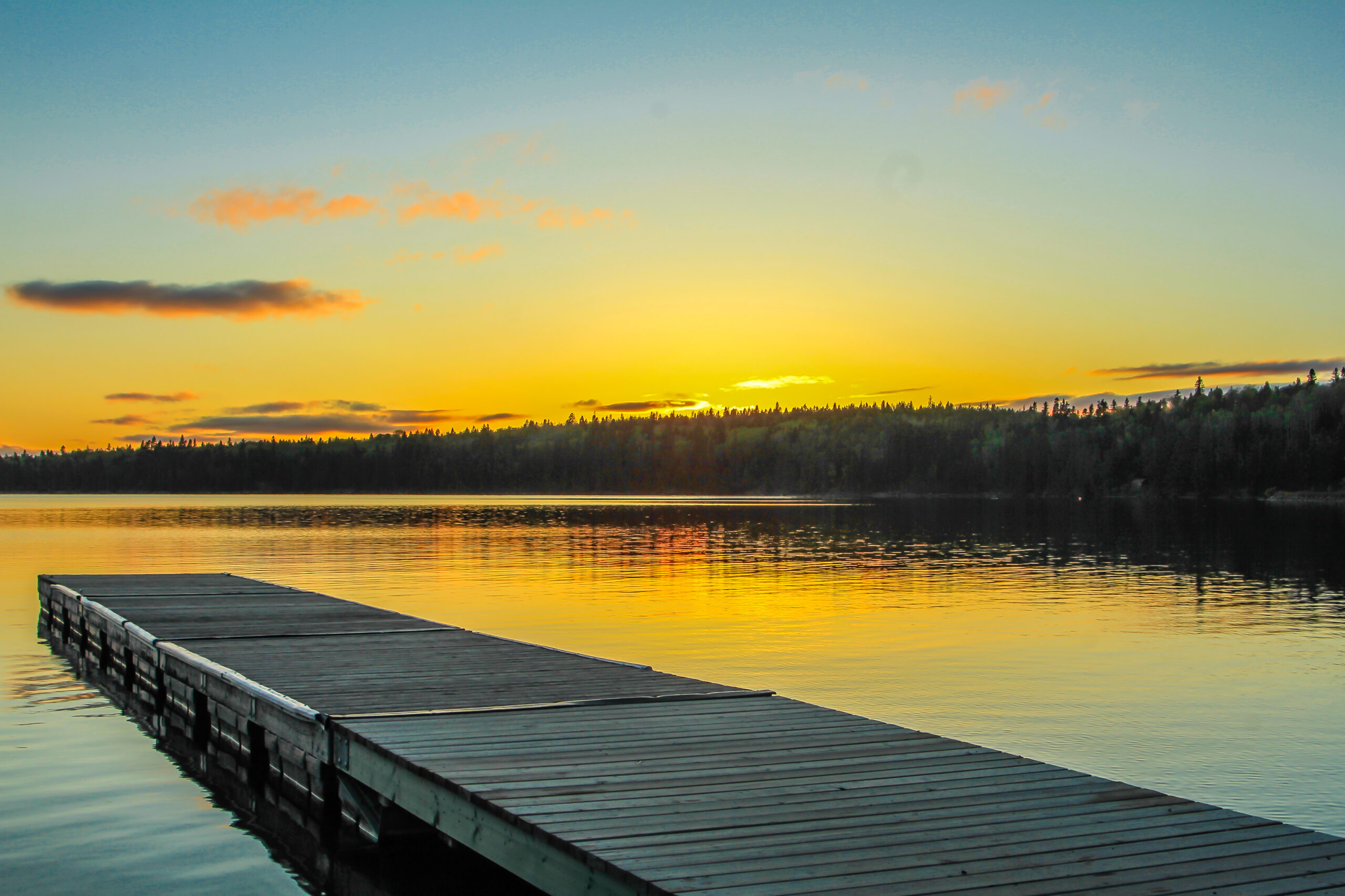 A wooden dock extends over calm lake water at sunset, perfect for Memorial Day boating, with trees along the shore and the sky glowing orange and yellow.