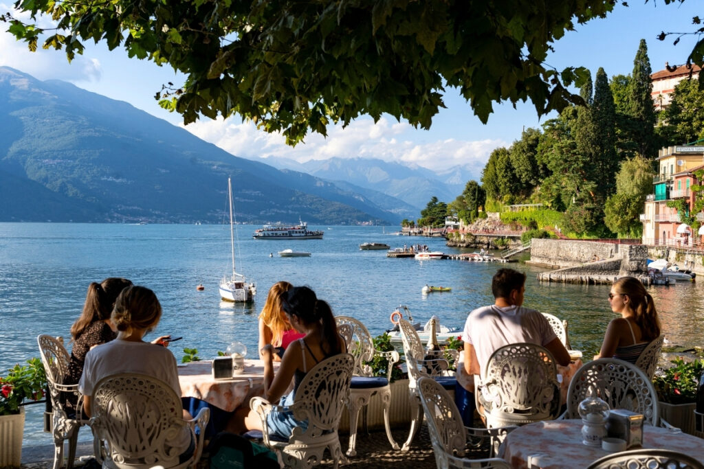People sitting at an outdoor cafe by a boating lake with boats, surrounded by mountains and greenery under a partly cloudy sky.