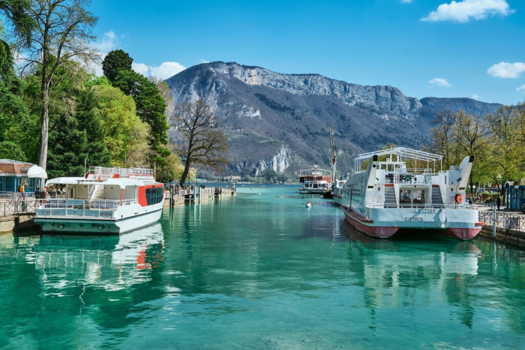 Two white boats are docked on a calm, turquoise boating lake surrounded by trees, with mountains and a clear blue sky in the background.