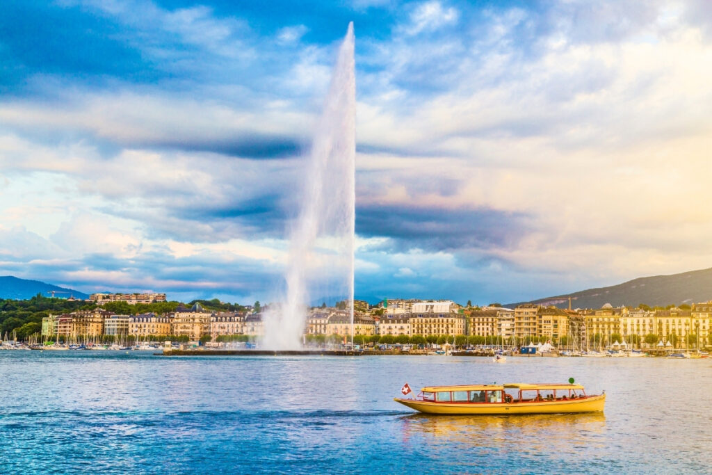 A yellow boat with a Swiss flag gliding across a boating lake, while the Jet d’Eau fountain sprays water high and buildings rise in the background under a partly cloudy sky.