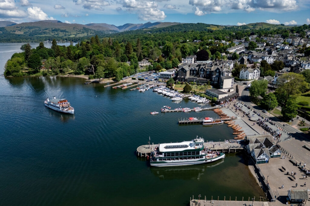An aerial view shows a lakeside town where boats are docked at piers on a boating lake. A ferry is gliding by, and green hills stretch in the background under a partly cloudy sky.