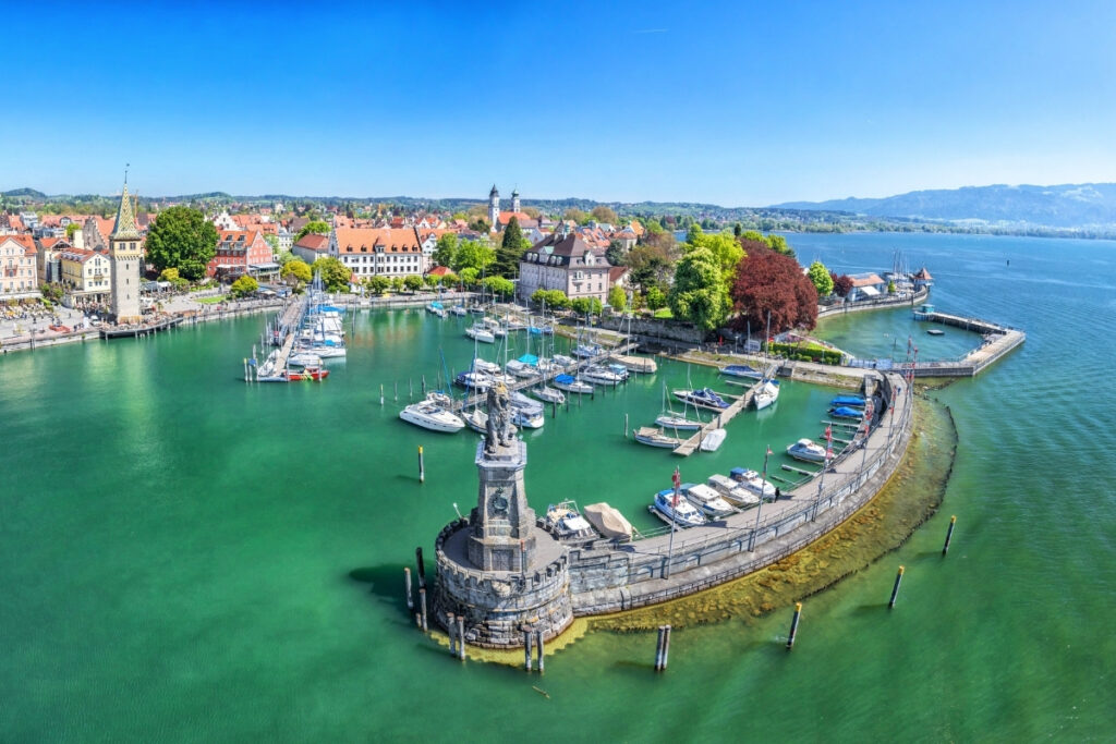 An aerial view of a boating lake marina with boats, a stone lighthouse tower, and historic buildings lining the waterfront under a clear blue sky.