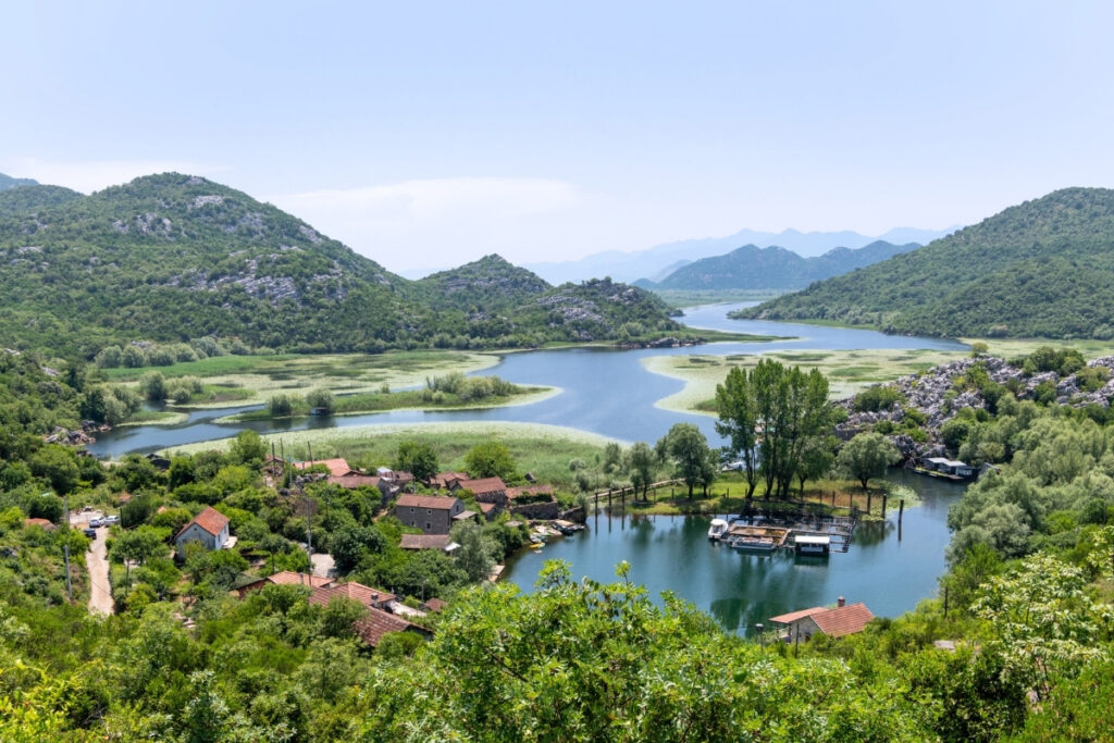 A small riverside village with red-roofed houses and boats sits among lush green hills by a calm boating lake under a clear sky.