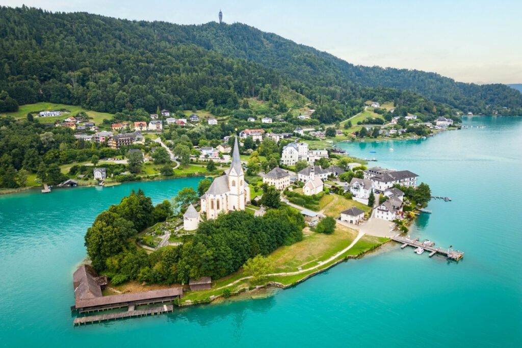 An aerial view of a small lakeside village with a central church, nestled by a boating lake and surrounded by turquoise water and forested hills.