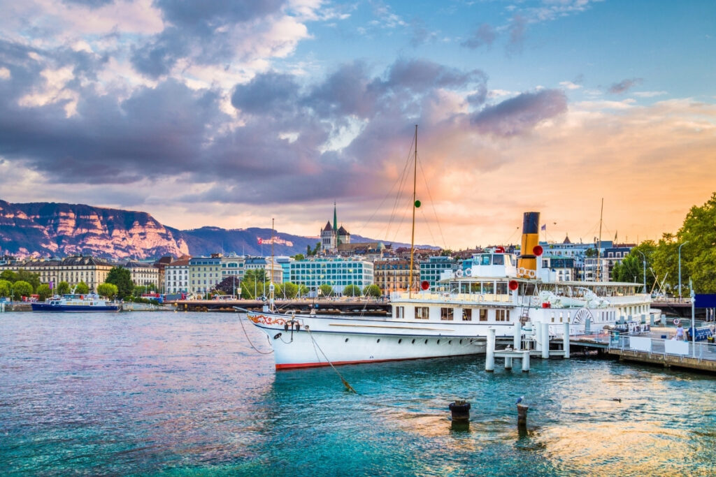 A white boat docked in a city harbor on a boating lake, with buildings, trees, and mountains silhouetted under a colorful sunset sky.
