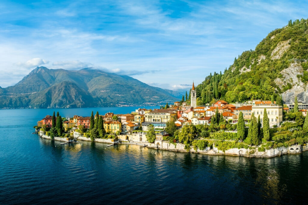 A scenic lakeside village with red-roofed buildings and lush greenery by a boating lake, set against mountains and blue water under a partly cloudy sky.