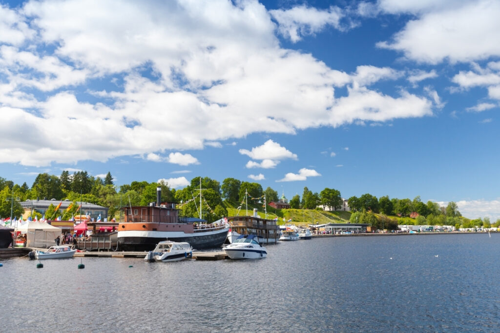 Boats are docked at a marina on a sunny day by the boating lake, with green trees, grass, and buildings in the background under a partly cloudy sky.