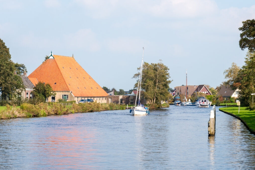 A sailboat gliding down a calm boating lake bordered by grassy banks and houses with orange roofs under a partly cloudy sky.