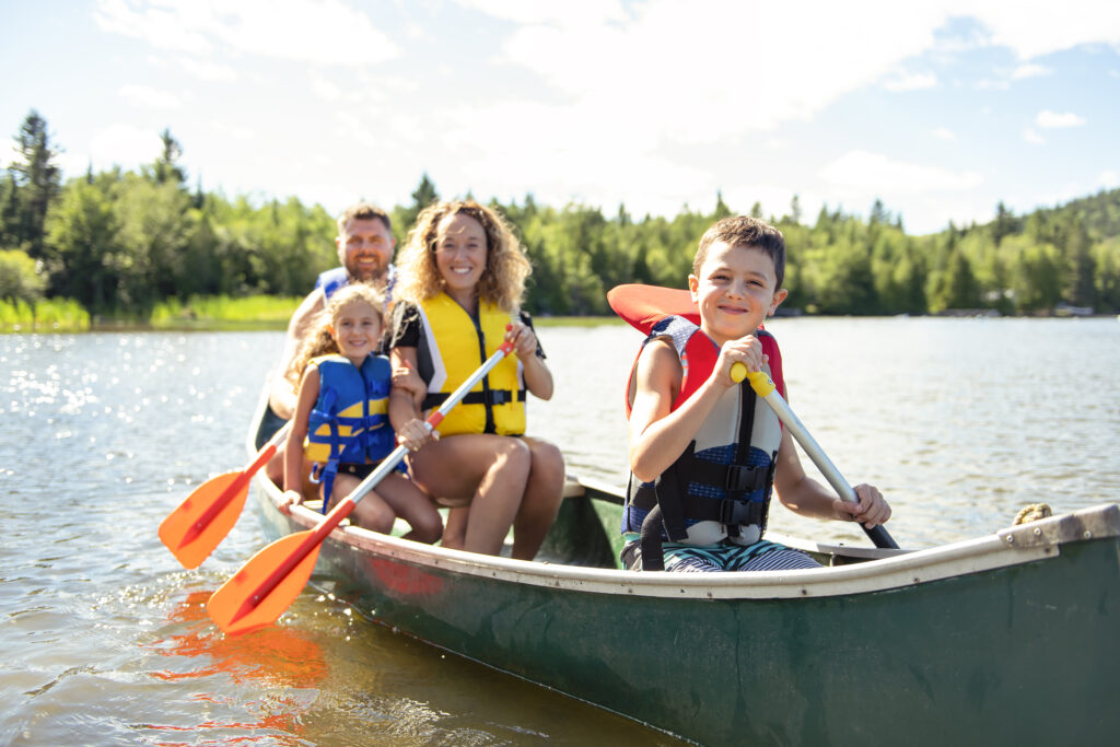 A family of four's wearing life jackets, making sure they are safe while boating, as they paddle a canoe on a lake, with trees and a blue sky in the background.