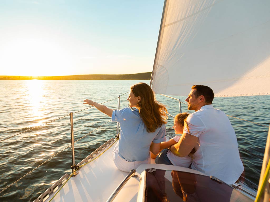 A family’s sitting together at the front of a sailboat, gazing out at the water and sunset under clear skies.