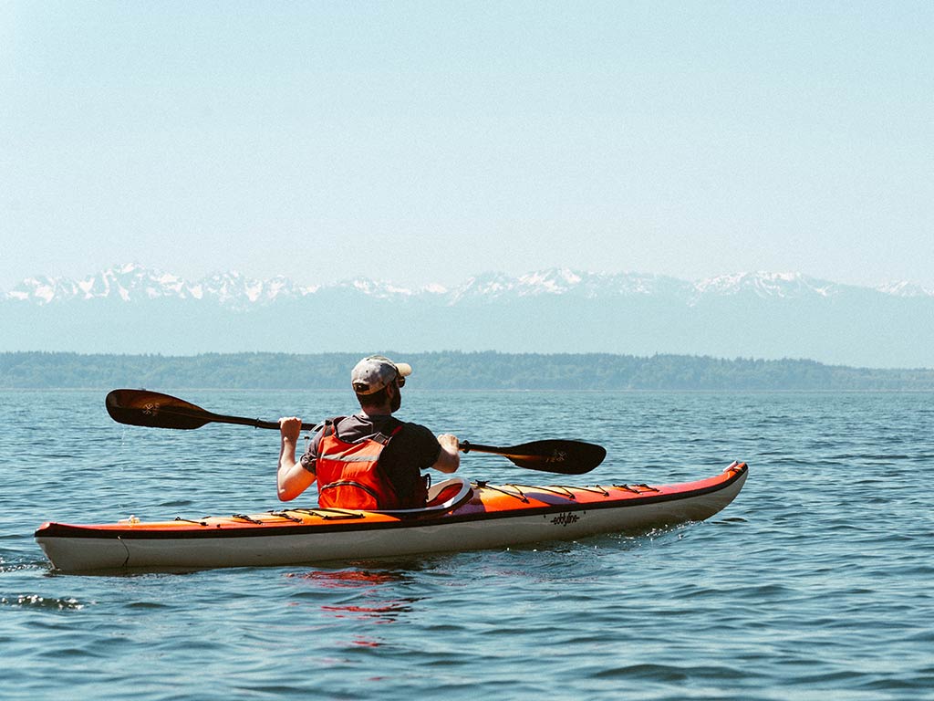 Someone's in a red kayak paddling on a calm body of water, with distant mountains on the horizon under a clear sky.