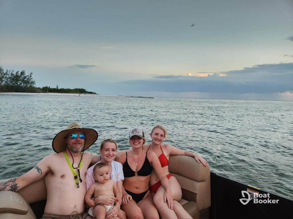Four adults and one baby are sitting on a pontoon boat with water and distant trees in the background under a cloudy sky.