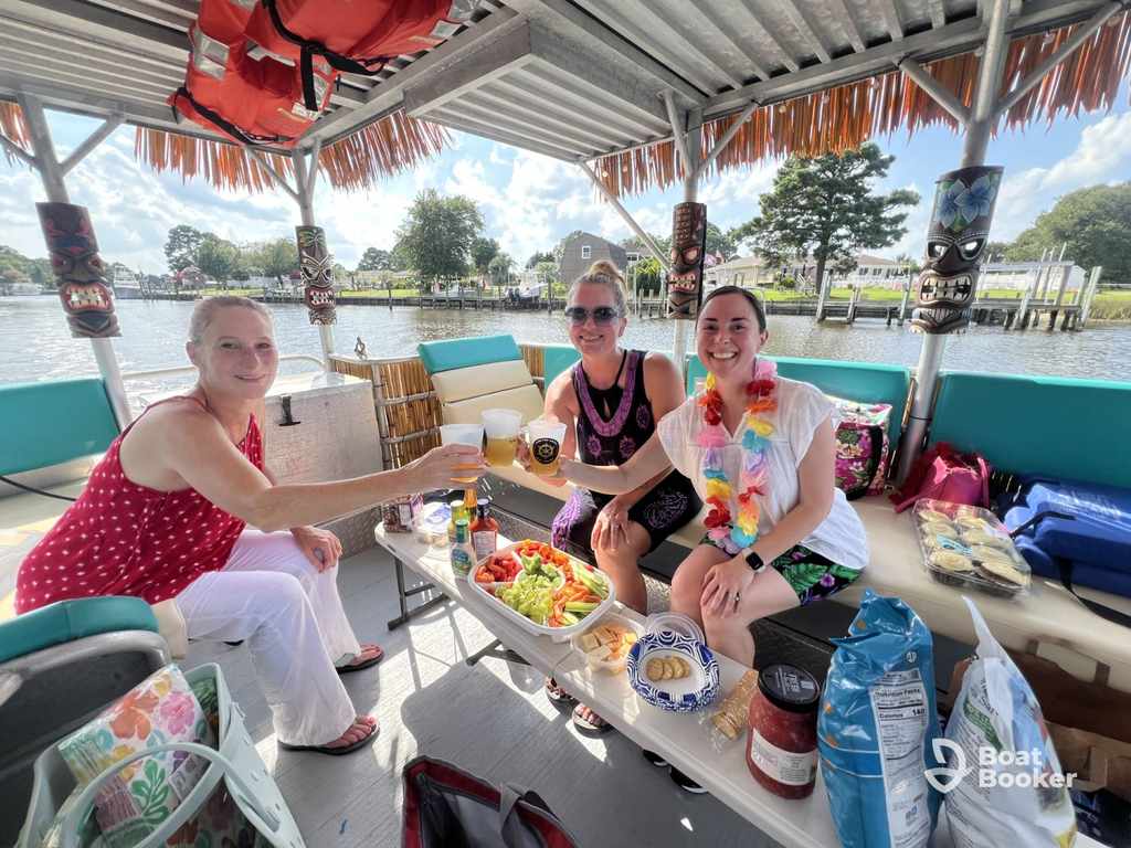 Three women sit around a table with food and drinks on a covered pontoon boat, enjoying a meal together on the water. Houses and trees are visible in the background.