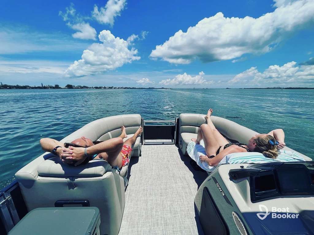 Two people are relaxing on loungers at the back of a pontoon boat out on calm water under a partly cloudy blue sky.