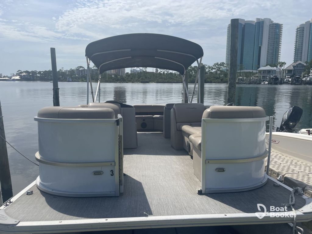 A pontoon boat with beige seating and a black canopy's docked on calm water, with tall buildings and trees in the background.