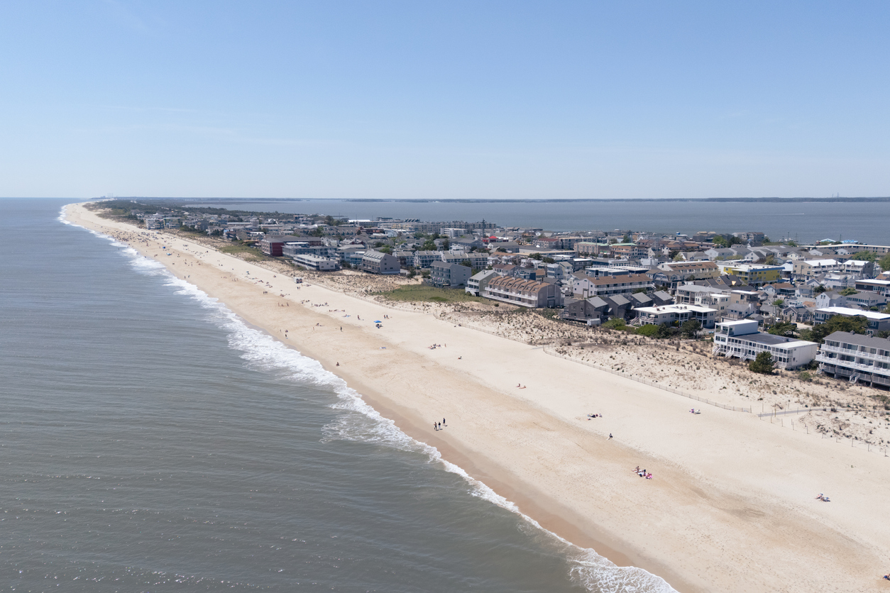 An aerial view of a sandy beach with waves on one side and a coastal town with houses and buildings on the other under a clear sky in Dewey Beach, Delaware.
