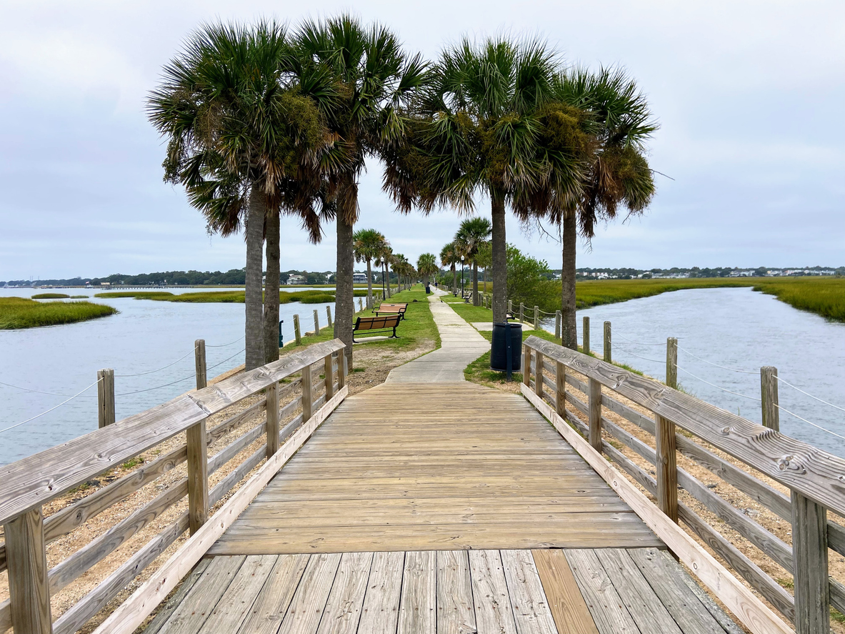 A wooden boardwalk with railings leads through palm trees along a waterfront path, surrounded by marshland and water under a cloudy sky in Mount Pleasant SC