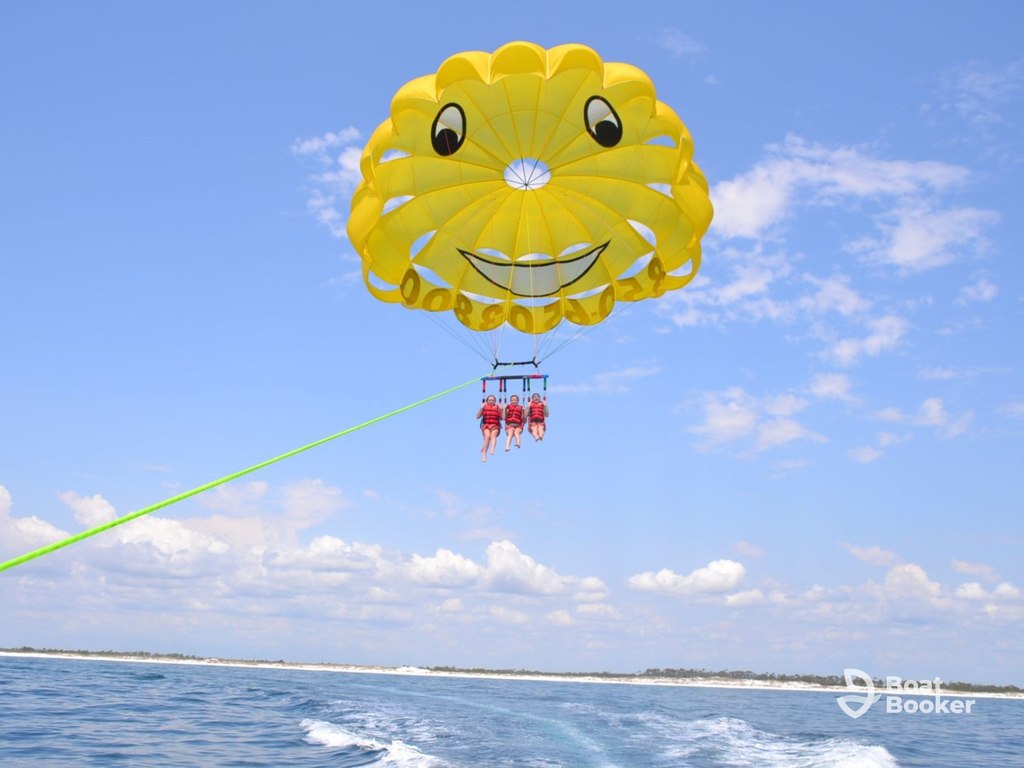 Three people are parasailing over the ocean, hanging from a yellow parachute with a huge smiley face. The sky's mostly clear with some scattered clouds, and the shoreline's visible far off.
