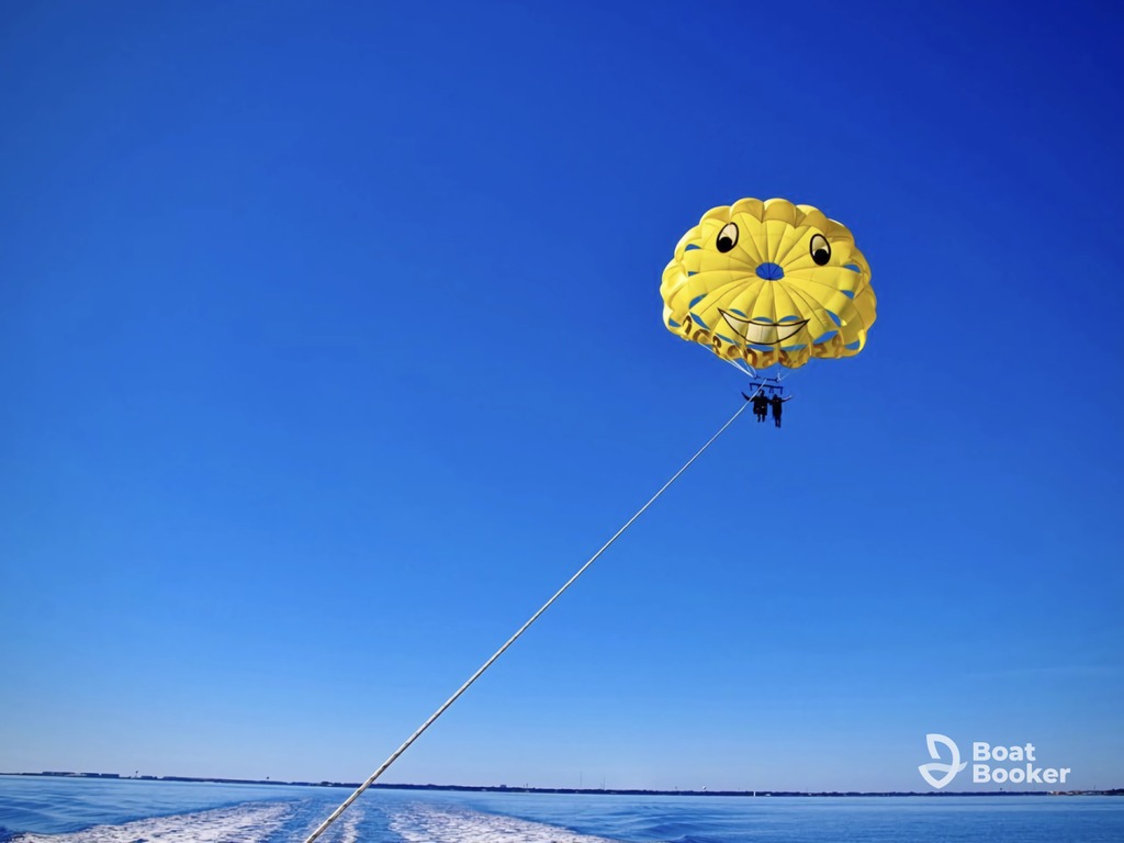 Two people are parasailing over the ocean, attached to a yellow parachute with a smiley face design against a clear blue sky. The Boat Booker logo's visible in the corner.