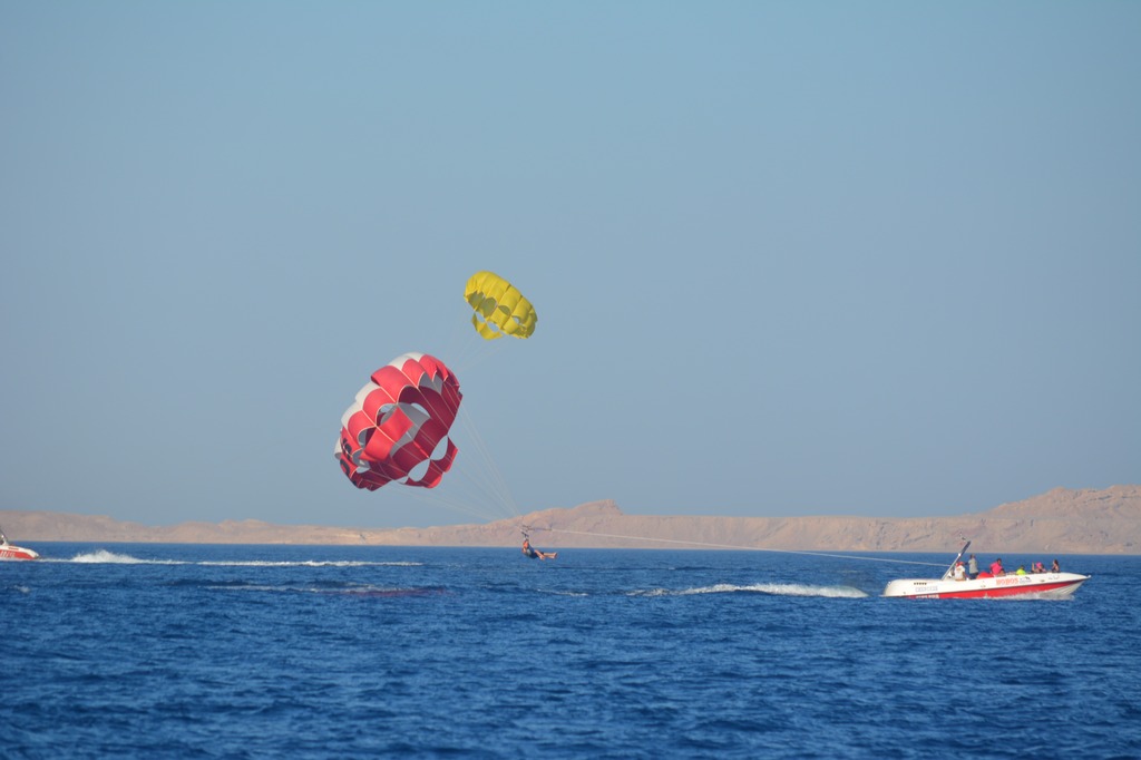 Two people are parasailing over the sea with bright Red and Yellow parachutes, each pulled by a speedboat, and a distant coastline's visible beneath a clear sky.