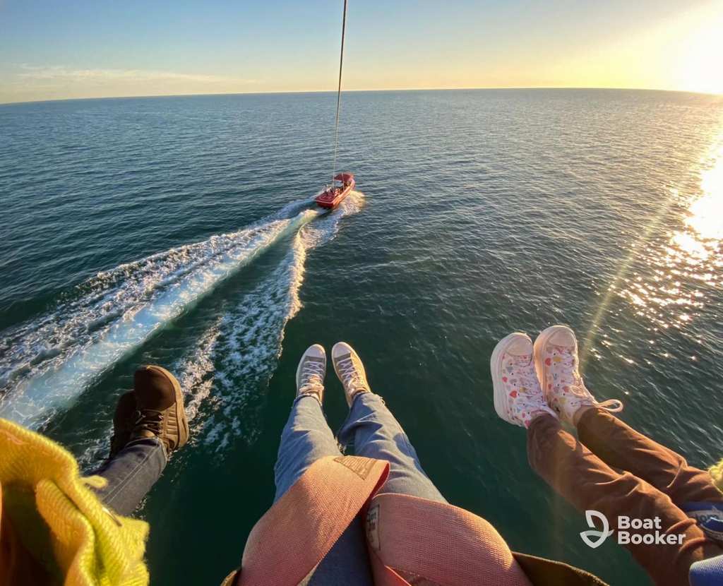 Three people are parasailing above the ocean, their feet visible, as a boat pulls them at sunset.