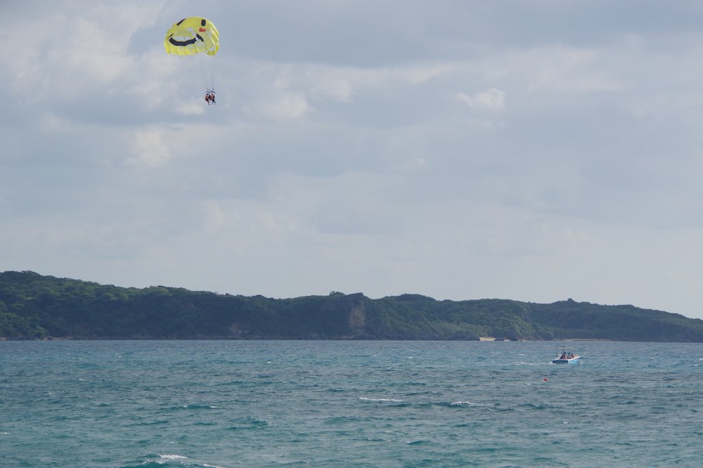 A person’s parasailing above the ocean, attached to a yellow parachute with a smiley face, while a boat tows them close to a forested coastline under a cloudy sky.