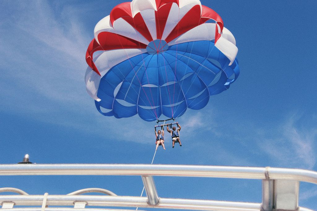 Two people are parasailing together, hanging from a red, white, and blue parachute in a clear blue sky above a metal boat railing. There aren't any FISH or specific species visible.