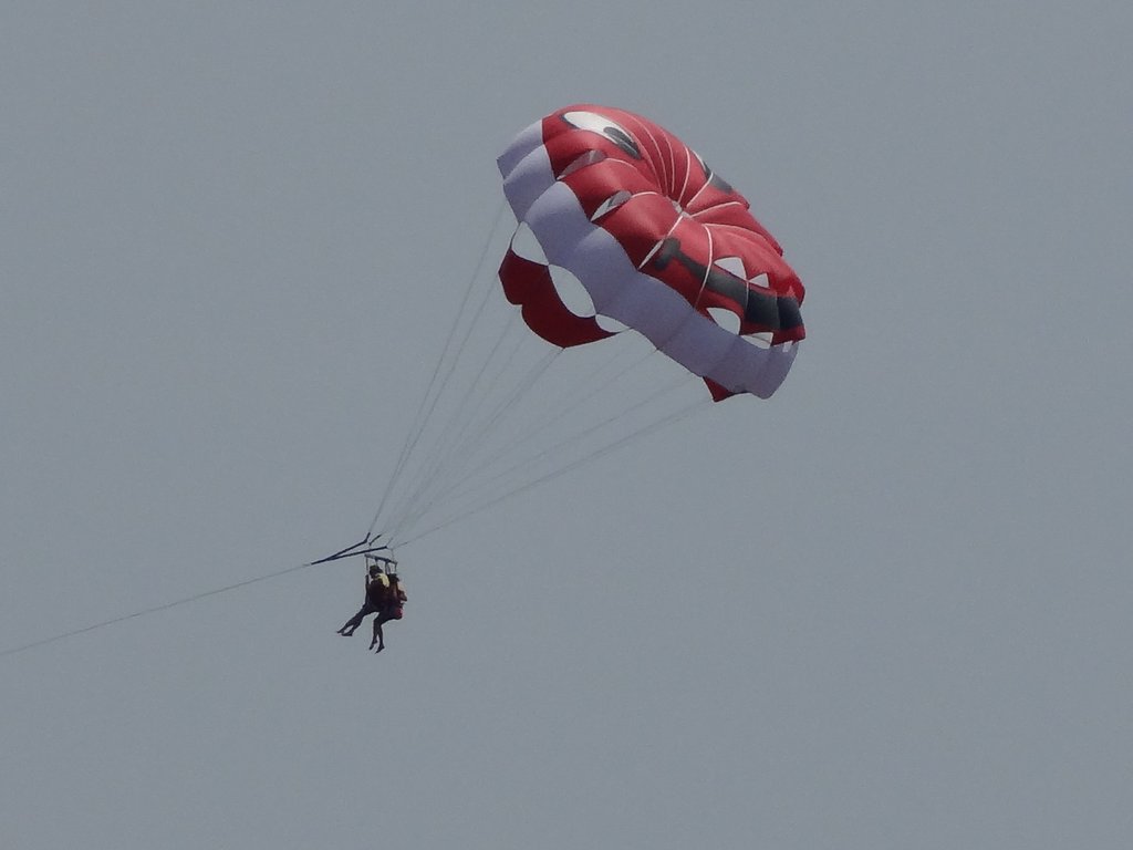 Two people are parasailing, suspended by a red and white parachute against a clear sky.