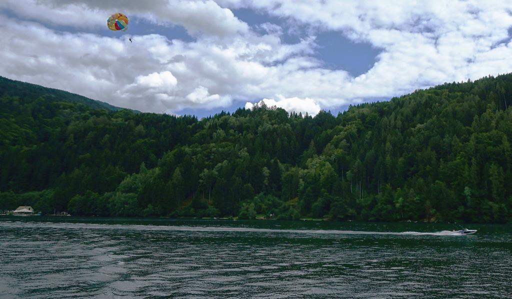 Someone's parasailing with a vibrant parachute over a lake, being towed by a speedboat; forested hills rise in the background beneath cloudy skies.