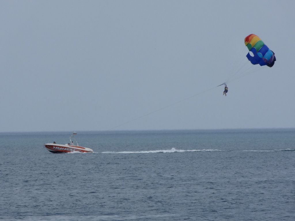 Someone's parasailing over the ocean, attached to a rainbow-colored parachute that's pulled by a red motorboat.