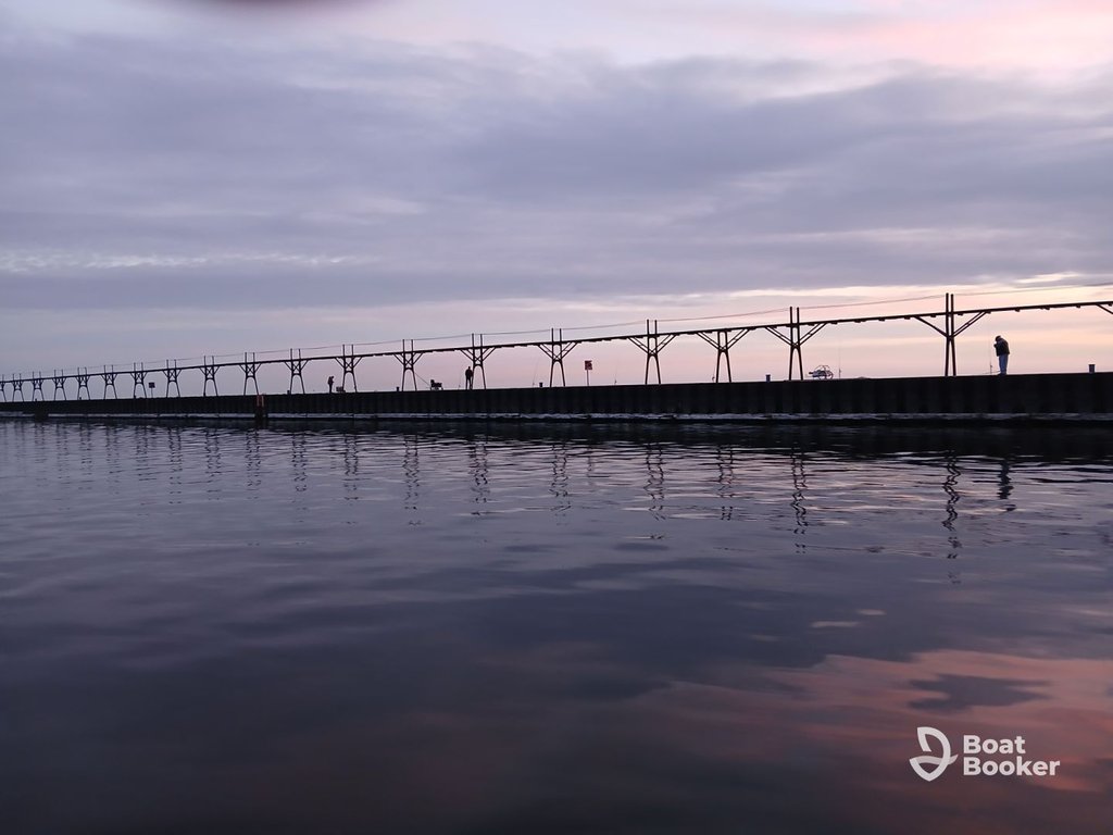 A long pier stretches over calm water at sunset, with several people standing and walking on it. The sky's overcast with scattered clouds. No Salmon or Trout are visible in the scene.