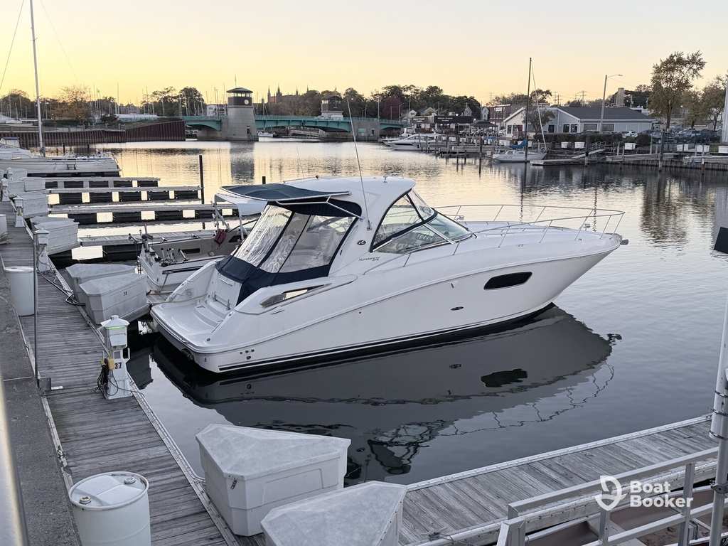 A white motor yacht's docked at a marina with calm water, empty docks, and buildings in the background as the sun sets.