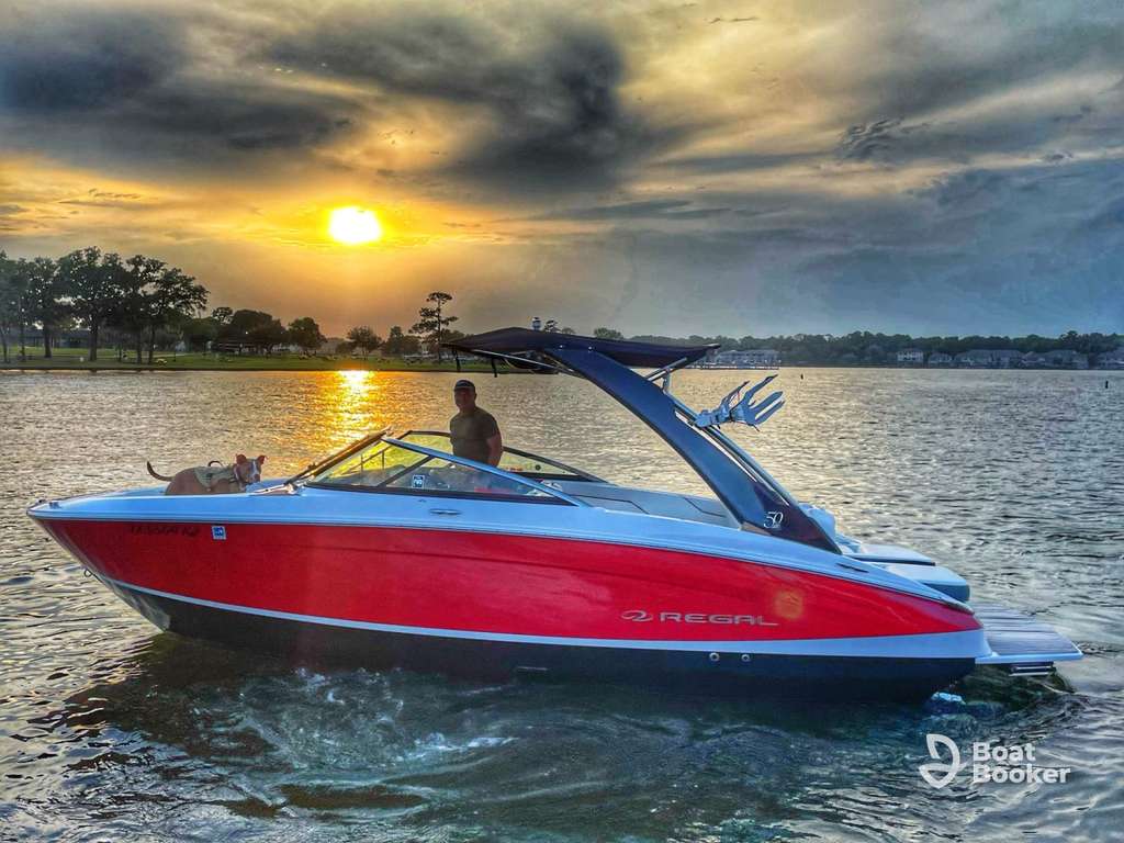 A red Regal speedboat with a man and a dog onboard floats on a lake at sunset, with trees and houses in the background.