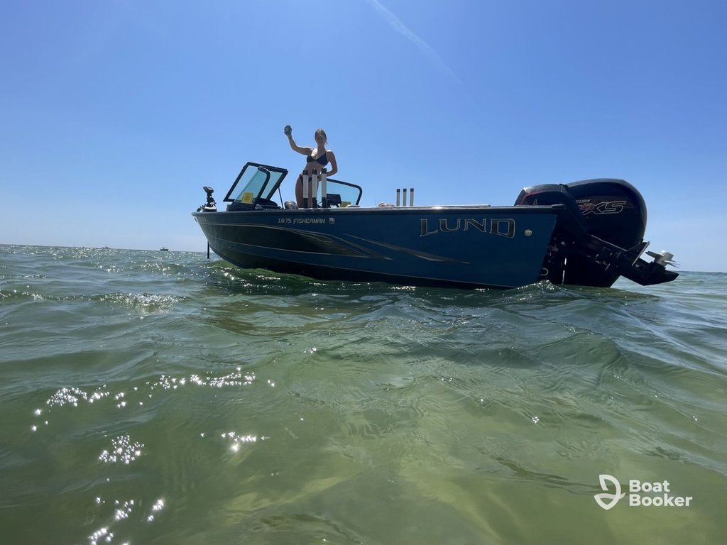 Someone's standing on a blue Lund fishing boat in calm, shallow water under clear skies. The "Boat Booker" logo's visible in the corner.