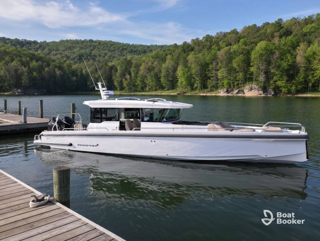 A white motorboat's docked near a wooden pier on a calm lake, surrounded by green, forested hills under a clear sky. The Boat Booker logo's in the lower right corner.