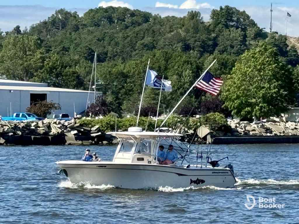 A small motorboat with three people onboard's cruising on a river, flying American and nautical flags, while trees and a building are visible on the shore.