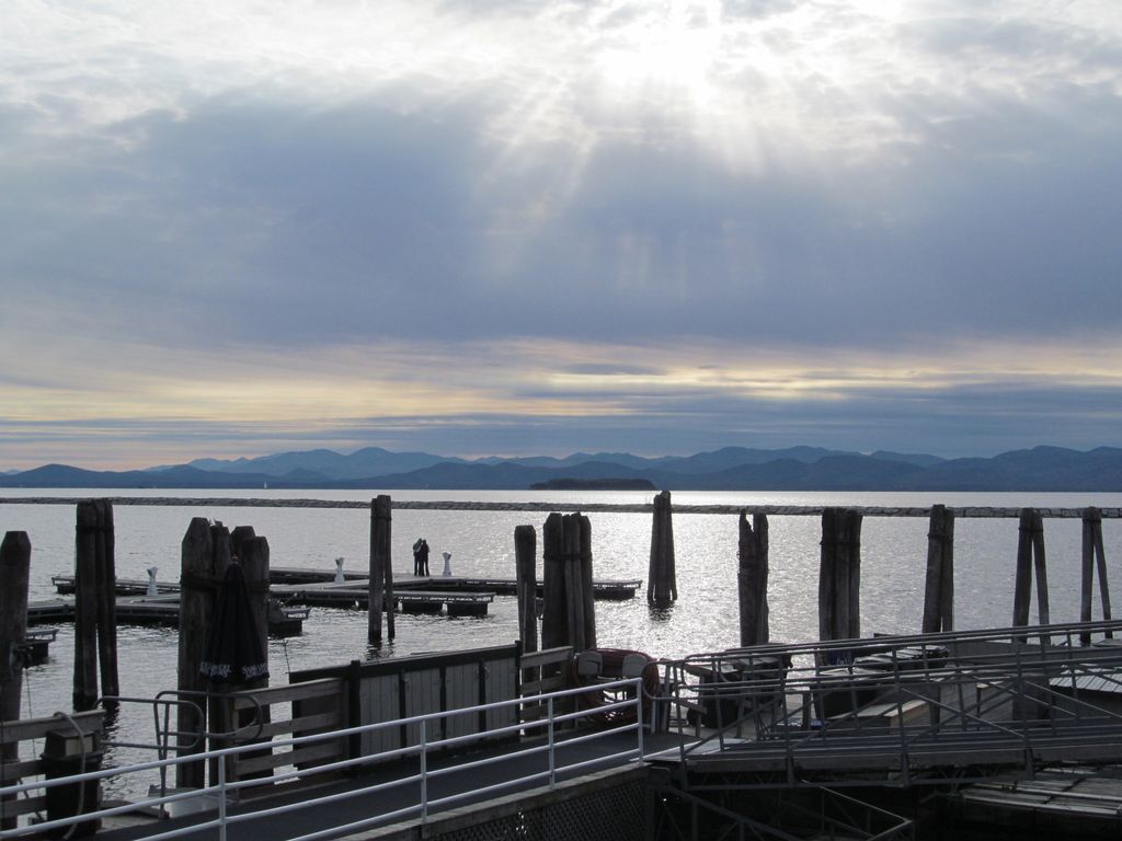 A waterfront dock with wooden posts stretches into a calm body of water, mountains in the back, and sunlight breaking through a cloudy sky. There aren't any fish or species visible here.
