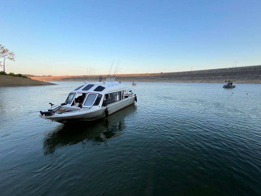 A white fishing boat with fishing rods is floating on a calm lake. You can see other boats in the distance and a shoreline in the background.