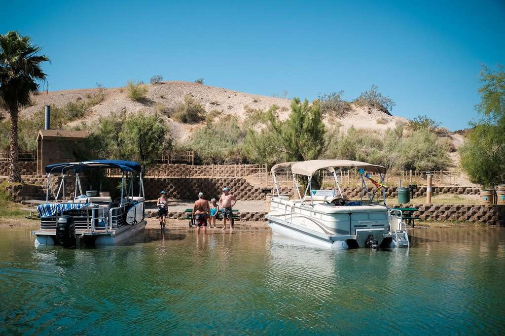 Two pontoon boats are docked by a sandy shore where a group of people stand near the water, with trees and a rocky hill in the background under a clear sky.