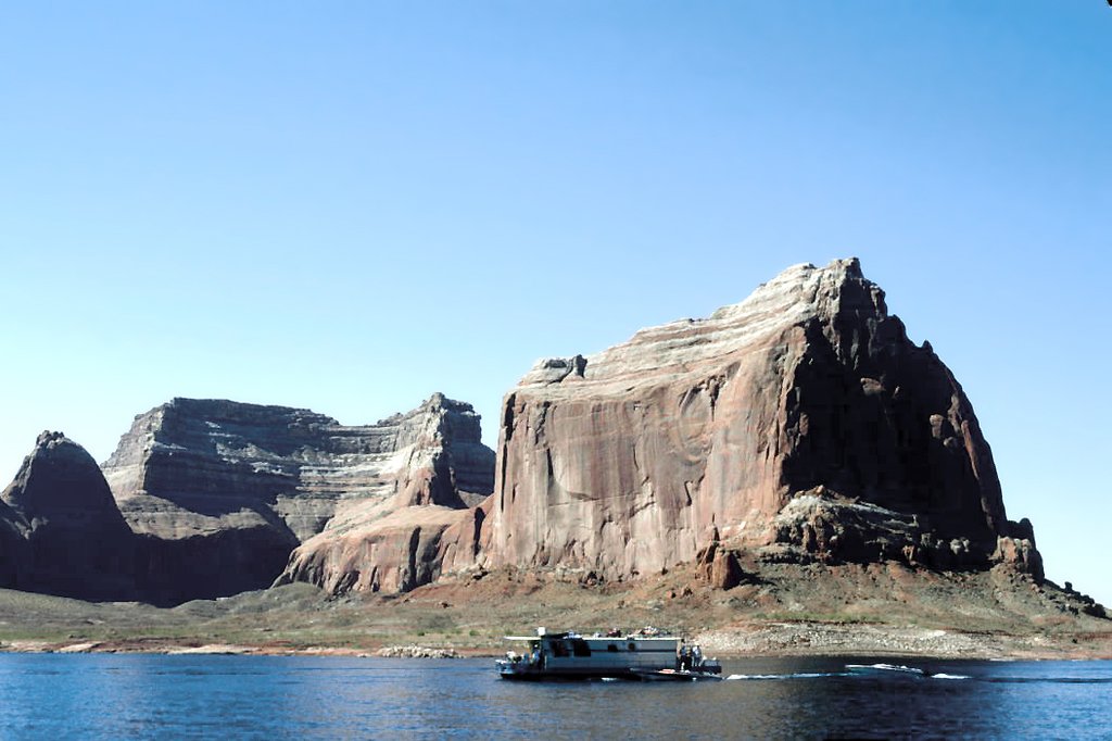 A houseboat's floating on a calm lake in front of large Red Rock formations under a clear blue sky.