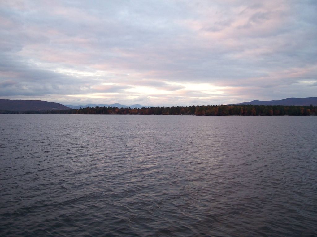 A calm lake with gentle ripples, bordered by a tree line and distant hills, sits under a cloudy sunset sky. There aren't any visible fish like Bass or Trout along the shoreline.