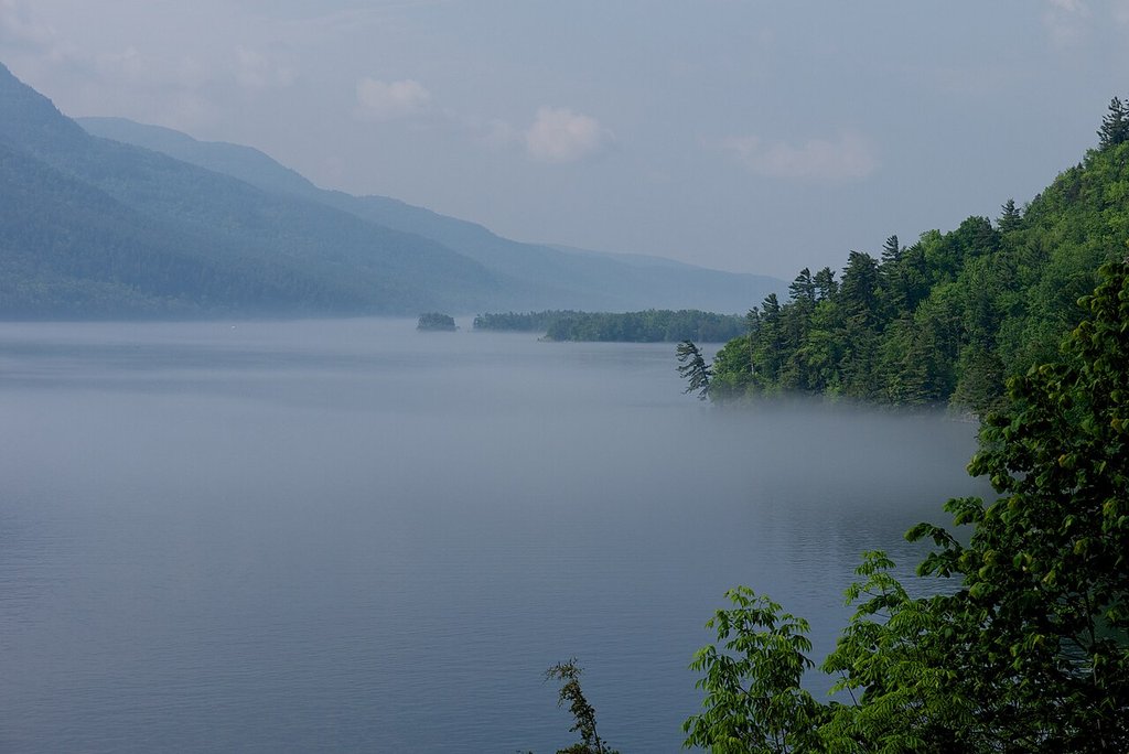 There's a calm lake with mist rising over the water, and it's bordered by forested hills and distant mountains beneath a cloudy sky.