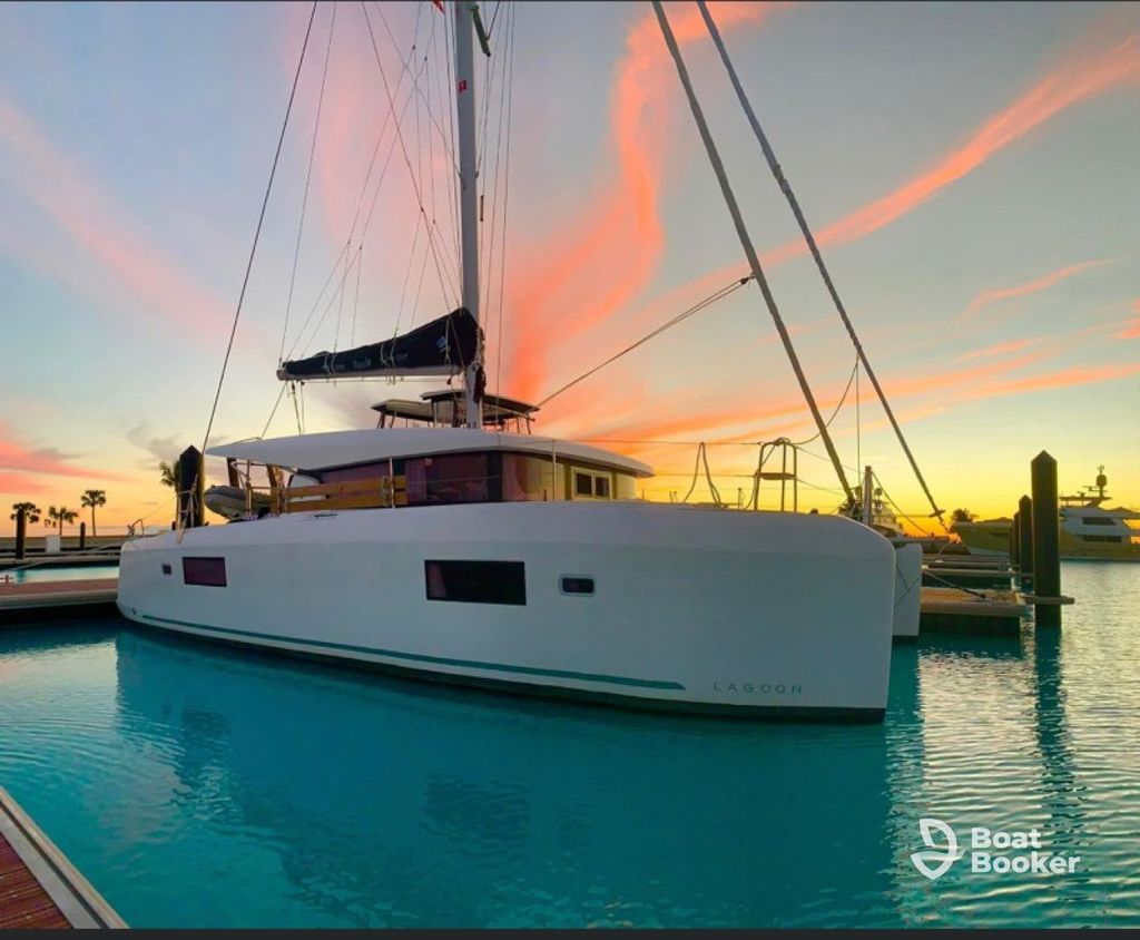 A white catamaran yacht's docked in a marina at sunset, with pink clouds in the sky and calm turquoise water. The "Boat Booker" logo's visible in the lower right corner.