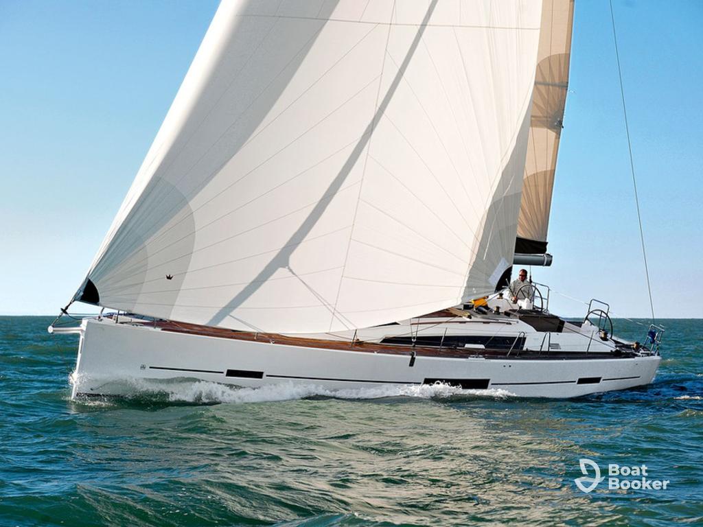 A white sailboat with large sails glides across the water on a sunny day, with two people visible on deck. The "Boat Booker" logo's in the lower right corner.