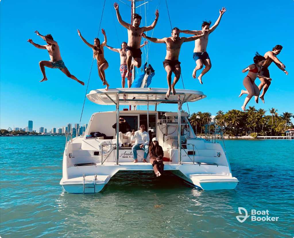 A group of people jump off the back of a white yacht into blue water, while others sit onboard. A city skyline and palm trees are in the background. No visible fish species in the scene.