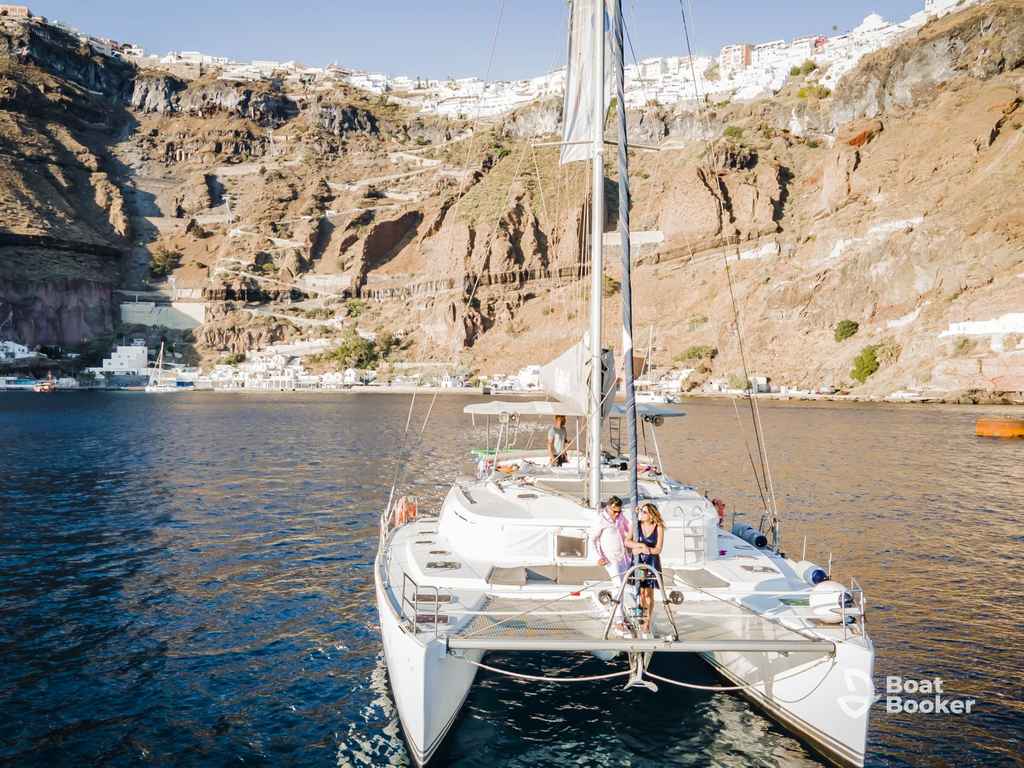 A white catamaran—it's a type of boat with two parallel hulls—is anchored near rocky cliffs and coastal buildings under a clear sky.