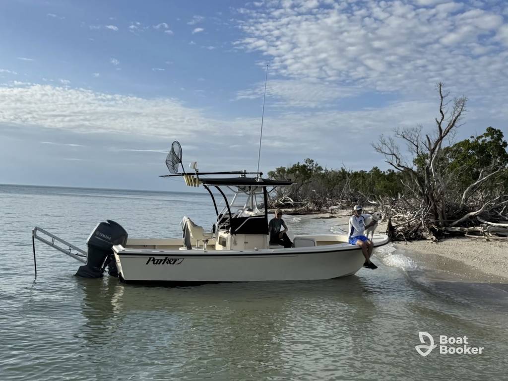 A small motorboat with two people on board's anchored near a sandy shore with trees and driftwood; "Parker" is visible on the boat's side.