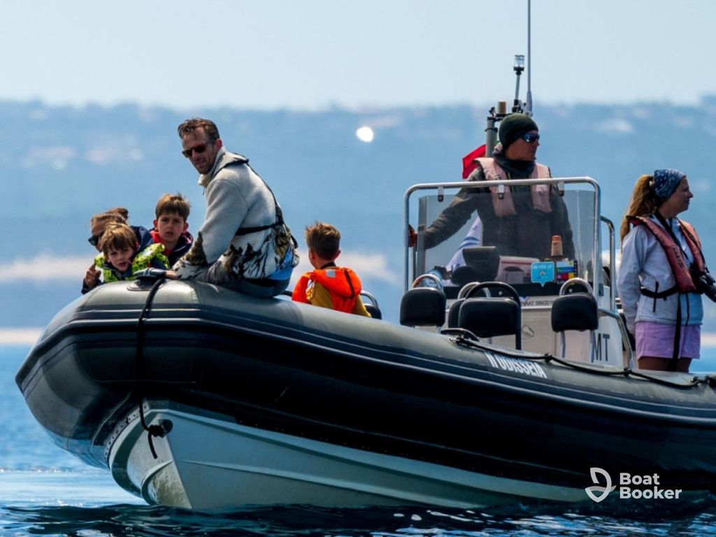 A group of adults and children in life jackets are seated on a black inflatable boat on calm water, with land visible in the background.